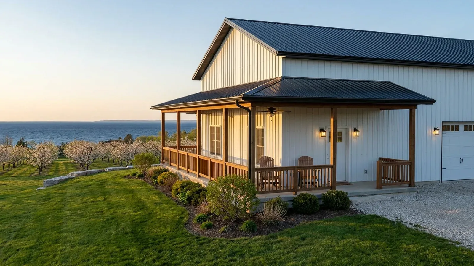 A white barndominium with a deep wrap-around covered porch beside a cherry orchard, open lake water on the horizon beyond.