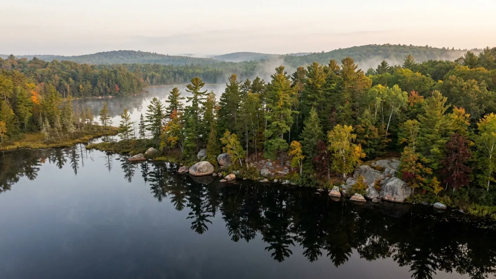 A still Northwoods lake ringed by tall white pines, birch and early autumn maples, with granite boulders on the shore.